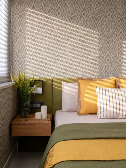 Sunlight streams through the blinds, casting beautiful shadows across the bed in the sunroom. The combination of Ikkat wallpaper, a floating wooden nightstand, and fresh flowers creates a warm and inviting atmosphere.