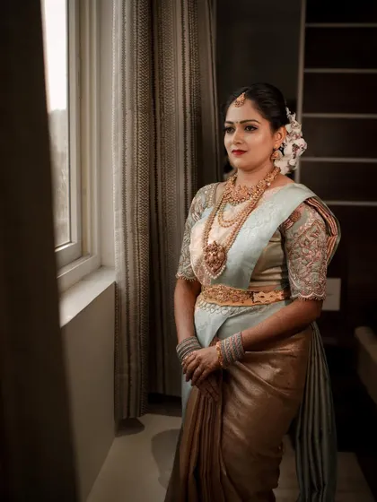 A pensive portrait of a bride in a beautiful two-tone saree, looking out a window. The lighting creates a soft, classic mood.