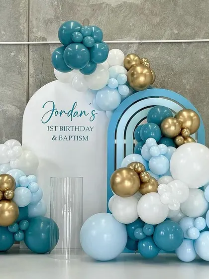 A modern and stylish balloon decoration for a first birthday and baptism. The setup uses arched backdrops and a sophisticated color palette of teal, blue, white, and gold.
