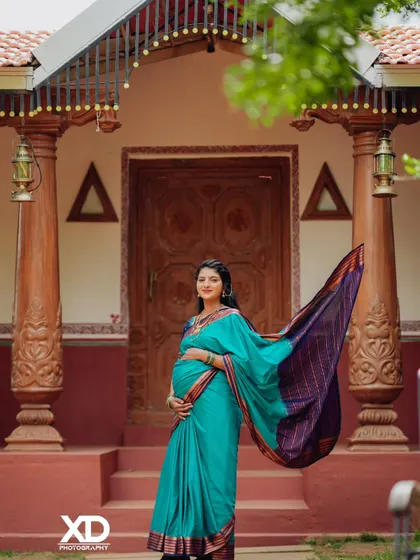 A stunning solo portrait of the mother-to-be. The flowing teal saree against the rustic backdrop of a traditional home creates a picture of pure elegance and grace.