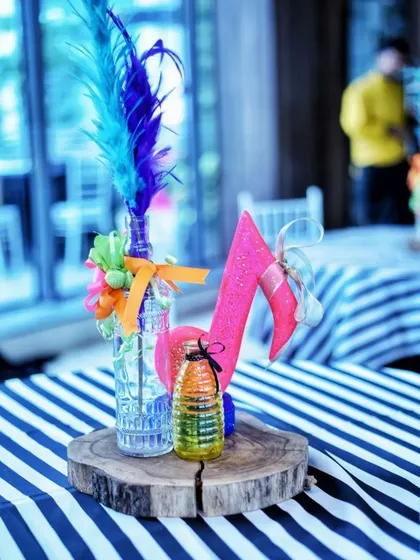 A close-up of a Rockstar party centerpiece, featuring a glass bottle with bright blue feathers and a sparkling pink musical note. The striped tablecloth adds to the bold, fun aesthetic.