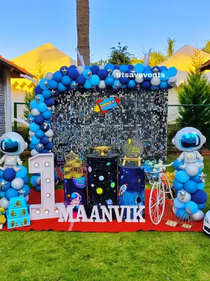 An out-of-this-world space theme for a first birthday, set up outdoors. This decoration includes a shimmering sequin wall, astronaut and rocket cutouts, and a large marquee number '1' to celebrate the big milestone.