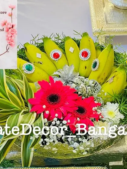 A bunch of fresh bananas decorated with kundan stones, arranged in a basket with bright red gerbera daisies and other foliage.