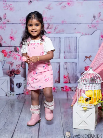 A little girl in a pink floral dress stands in a rustic, shabby-chic setting with a birdcage and teepee. The soft colors and charming props create a very sweet and gentle atmosphere.