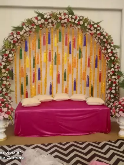 An indoor floral arch with colorful tassels hanging against a white backdrop. This is a great way to add a pop of color to your Mehndi or Haldi decor.