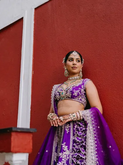 A bride in a bold purple lehenga poses against a contrasting red wall, creating a powerful and fashionable bridal portrait.
