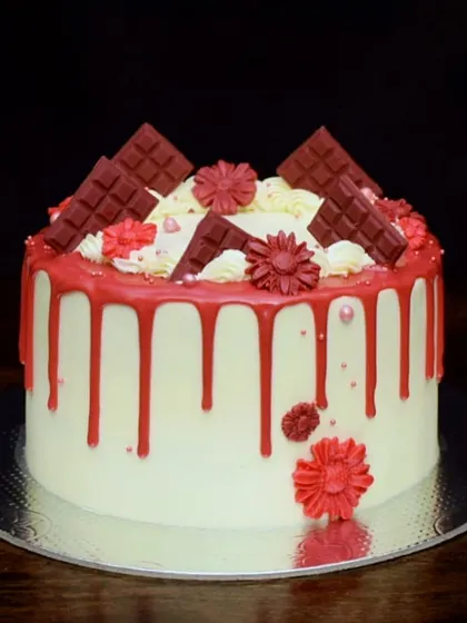 A red velvet drip cake, perfect for any celebration. This design features a vibrant red drip over a cream cheese frosting, topped with red fondant flowers and chocolate bars.