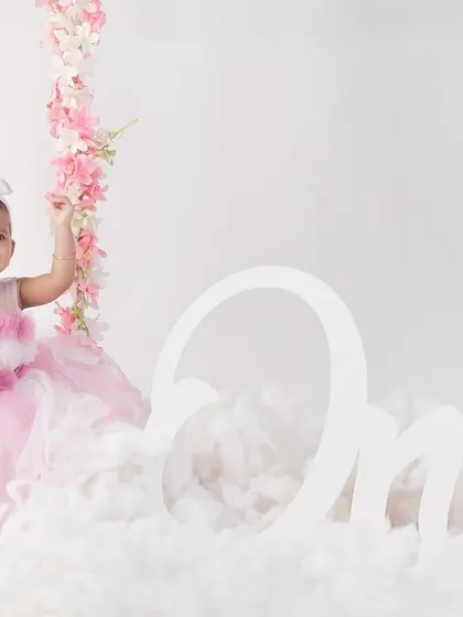 A beautiful and minimalist first birthday shoot. The simple white background, pink floral swing, and fluffy clouds keep the focus on the birthday girl's happy expression.