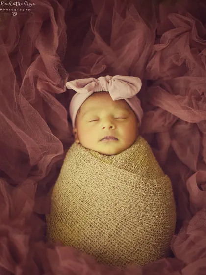 This 14-day-old doll is sleeping peacefully, wrapped in a rustic burlap-style swaddle and a pink bow headband.