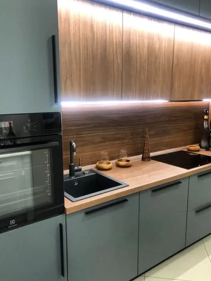 This kitchen combines matte grey cabinets with a warm wood-finish countertop and backsplash. The black handles, sink, and built-in oven provide a striking modern contrast.