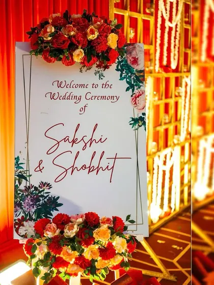A beautiful welcome sign for Sakshi and Shobhit's wedding ceremony. The sign is adorned with lush red and orange floral arrangements, perfectly matching the grand, traditional theme of the event.