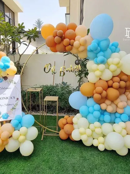 This outdoor baby shower decoration uses a gold ring backdrop and a vibrant balloon garland in orange, blue, and cream. A personalized welcome sign completes this cheerful and modern setup.