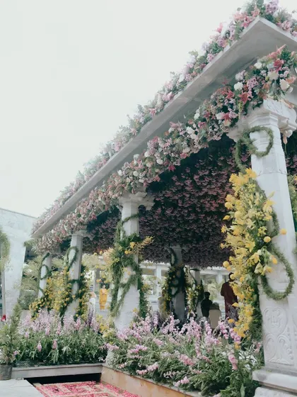 A closer look at the floral canopy from Nithya and Payton's wedding. The structure is covered in a dense arrangement of pink and yellow flowers, creating a stunning visual against the stone pillars.