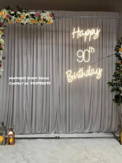 An elegant and simple setup for a 90th birthday. It features a white curtain backdrop, a 'Happy 90th Birthday' neon sign, and minimal floral arrangements with lanterns for a classic look.