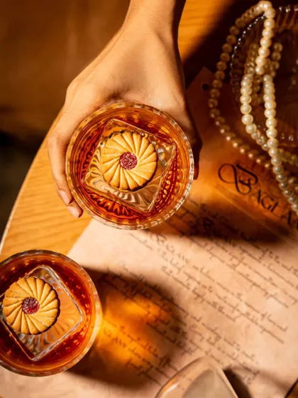 An overhead shot of the biscuit-garnished cocktail at Baglami. This perspective highlights the unique presentation and the rich color of the drink against the textured table and vintage-style menu.