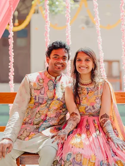 A smiling couple at their Mehendi ceremony, seated on a swing decorated with pink drapes and floral garlands.