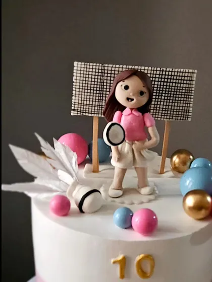 A badminton theme cake for a 10th birthday. The design includes a handmade figurine, an edible shuttlecock made with wafer paper, and a pink, white, and blue color scheme.
