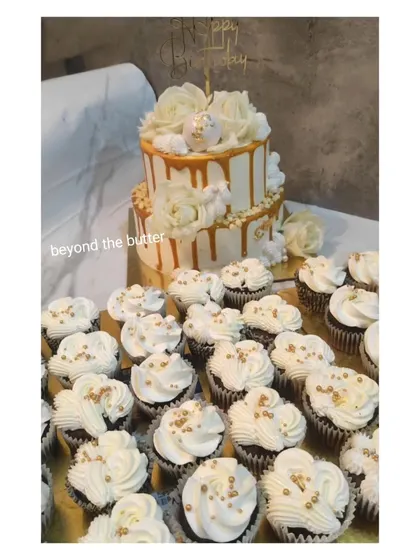 An elegant white and gold drip cake paired with matching cupcakes. The two tier birthday cake is topped with white roses and the cupcakes are sprinkled with gold beads, creating a coordinated and luxurious dessert table.