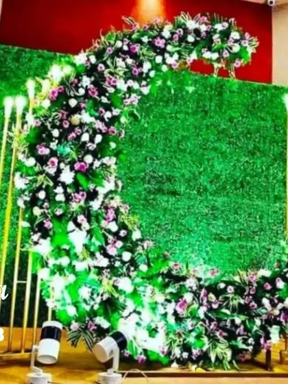 A modern and stylish naming ceremony photo point. A large floral crescent is set against a green hedge wall, flanked by tall, glowing candle stands for a dramatic and chic look.