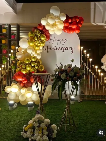 An outdoor anniversary decoration featuring a white arched backdrop with custom text. The backdrop is framed by a balloon garland in red, gold, and cream, perfect for a garden or terrace party.