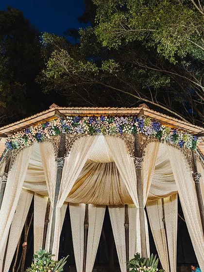 An evening view of a Sangeet mandap, elegantly draped in white and accented with purple and blue florals. The subtle lighting creates a sophisticated and dreamy atmosphere for the night's performances.