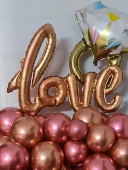 A close-up of a balloon arrangement featuring rose gold and pink balloons, topped with a "Love" foil balloon and a diamond ring balloon. This is a perfect centerpiece for an engagement or anniversary party.