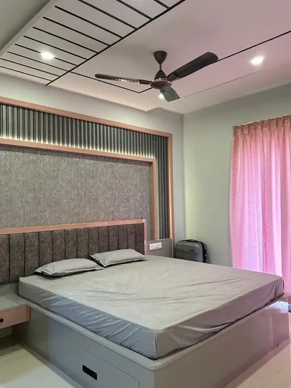 A cozy modern bedroom featuring a grooved false ceiling and exposed rafters. The grey wallpaper and upholstered headboard add layers of texture and warmth.