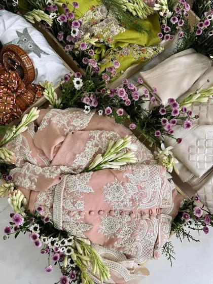 A close-up of a pastel pink outfit in a wooden tray, surrounded by fresh tuberoses and purple daisies. The fresh flower decoration adds a special touch.