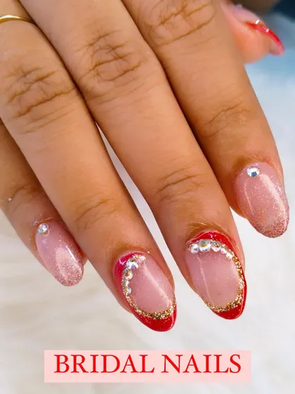A close-up of a glamorous bridal nail design, featuring a sheer glitter base with a red and gold glitter French tip, framed with rhinestones.