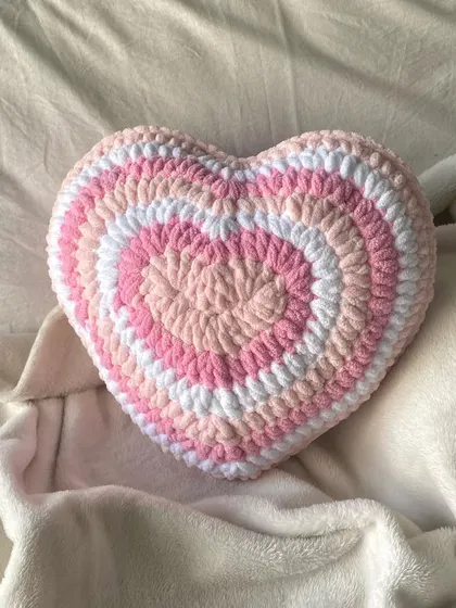 Our crochet heart pillow sitting on a soft, neutral blanket. The y2k-inspired design and colors are fully customizable to match your aesthetic.
