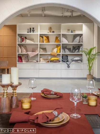 A view across a set dining table towards a large, built-in shelving unit displaying a curated selection of cushions. This layered view demonstrates how the store is designed to create beautiful compositions from every angle.