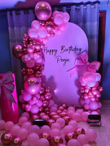 A beautiful pink-themed birthday party for Pooja. The setup includes a custom arch backdrop, an elaborate balloon garland with chrome accents, and decorative bow props.