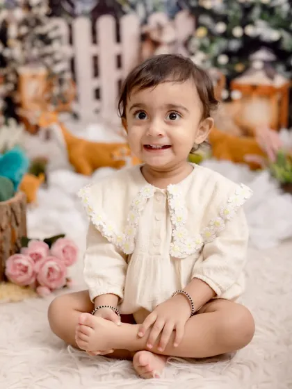 A beautiful sitter session portrait capturing a delightful, expressive smile. The baby is dressed in a lovely cream-colored romper, sitting in a soft, whimsical floral setting.