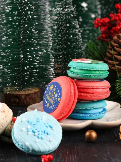 A close-up of colorful Christmas macarons, stacked against a backdrop of miniature pine trees and festive decor.