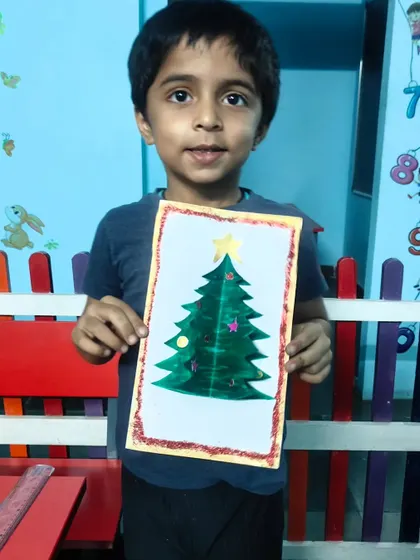 A student holds up the beautiful Christmas tree greeting card he made, decorated with glitter and a star.