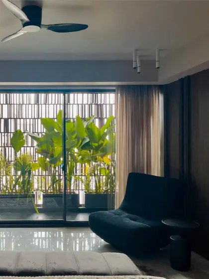 A serene bedroom corner where black metal and glass slats filter daylight while maintaining a visual connection to the balcony greenery. The sheer curtains and modern lounge chair create a perfect spot for quiet relaxation.