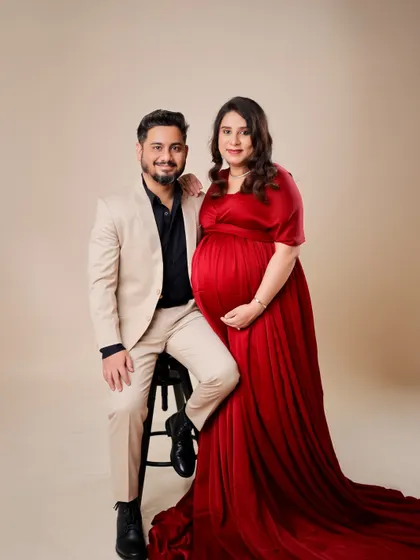 An elegant studio portrait of an expecting couple. The woman's flowing red gown and her partner's coordinated suit create a timeless and sophisticated image.