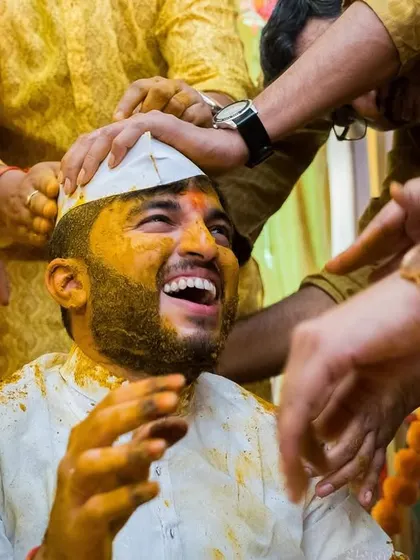 The groom bursts into laughter as his friends and family enthusiastically apply haldi.
