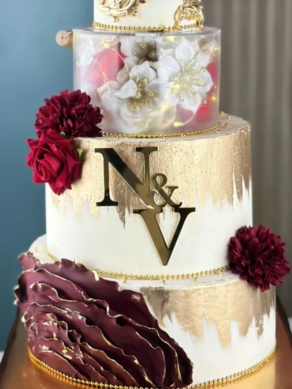 A touch of drama, a whole lot of elegance. This tall, four-tiered cake combines a rich maroon with brushed gold, featuring a clear tier filled with glowing lights and handcrafted flowers.