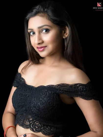 A classic studio portrait against a black background. The model wears a black lace off-shoulder top, and the soft lighting creates a gentle and inviting mood.