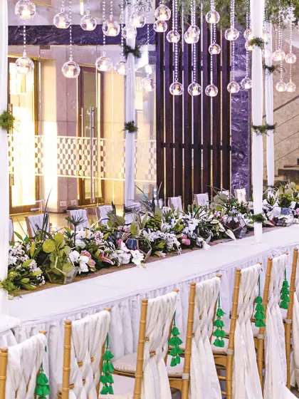An elegant long table setup with white roses, lilies, and hanging glass baubles. The green tassels on the chairs add a subtle pop of color and a unique, customized touch to the decor.