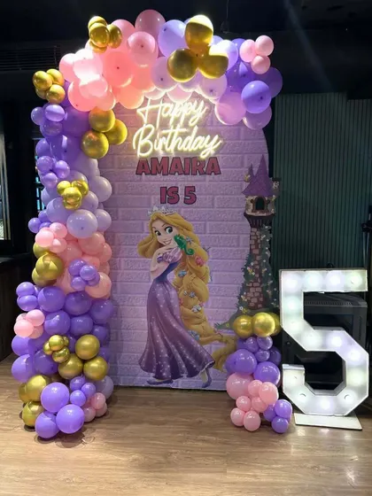 A Rapunzel-themed fifth birthday party. The arched backdrop features the princess against a brick tower background, framed by a purple, pink, and gold balloon garland.