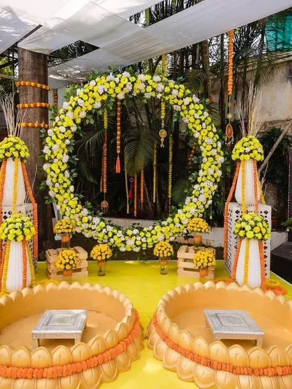 An elaborate outdoor Haldi setup with two 'urli' stations for the bride and groom. The backdrop is a large circular floral wreath made of yellow and white flowers.