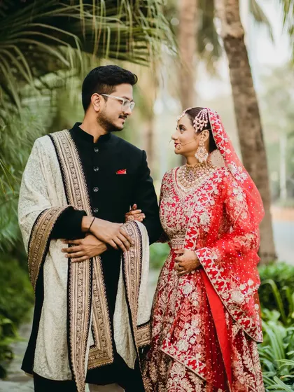 A classic portrait of a bride and groom, where her vibrant red lehenga stands out against the lush greenery. The way they look at each other speaks volumes about their love story.