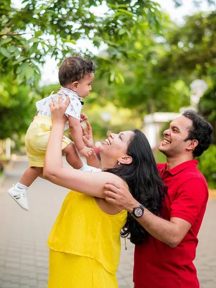 A happy family of three enjoying a moment in the park, with the mother lifting their baby high. We focus on capturing natural light and joyful interactions.
