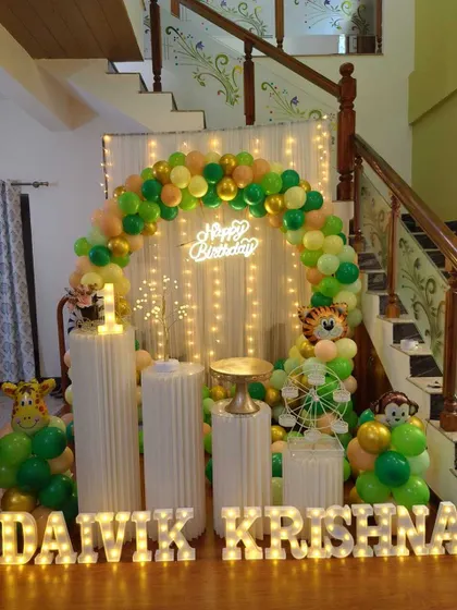 A jungle-themed birthday party set up near a staircase. The decor includes a green and gold balloon arch, a fairy light curtain, and animal props.