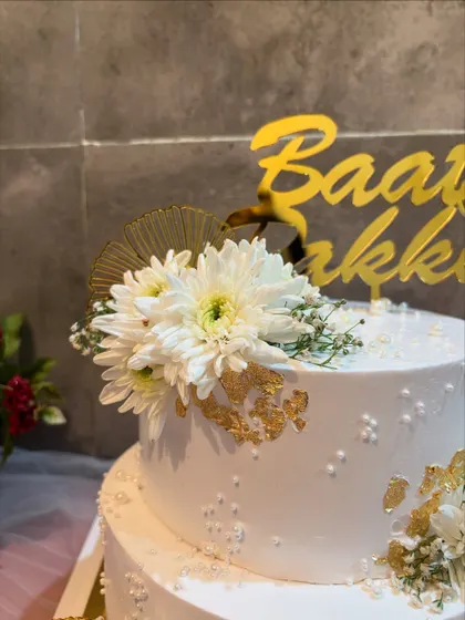 A close-up view of a two-tier Baat Pakki cake, showing the detail of the fresh white flowers, gold leaf, and delicate pearl sprinkles.
