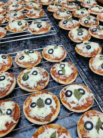 A batch of our freshly baked mini pizzas cooling on a wire rack. Each one is topped with tomato sauce, mozzarella cheese, a black olive, and a basil leaf.