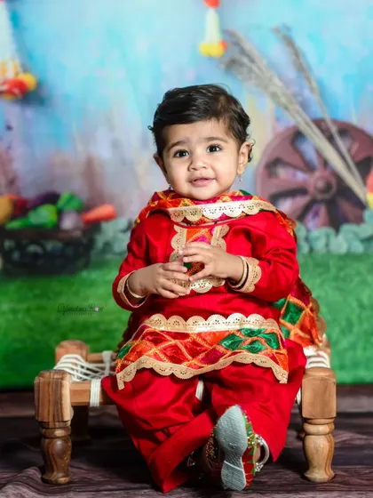 Celebrating Lohri, Pongal, or Makar Sankranti with a festive photoshoot. This little one is dressed in a vibrant red Punjabi suit, sitting on a traditional charpai.