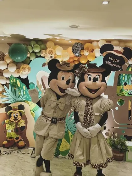 Mickey and Minnie mascots posing in front of a vibrant jungle-themed photo wall, complete with a balloon garland and character cutouts.